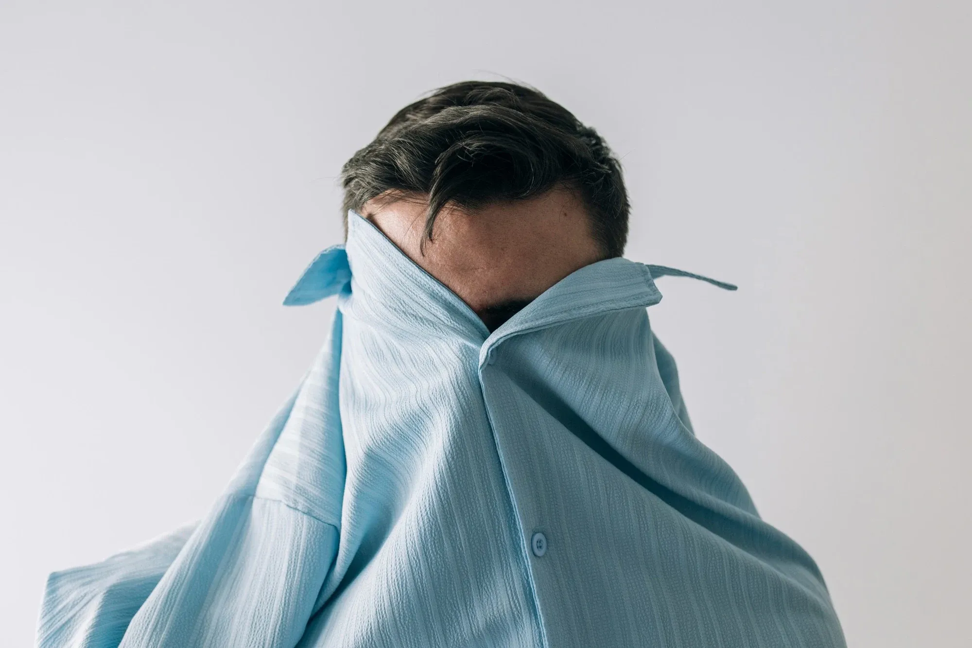 A portrait of a person in a pale blue shirt, their face partly hidden by its collar.