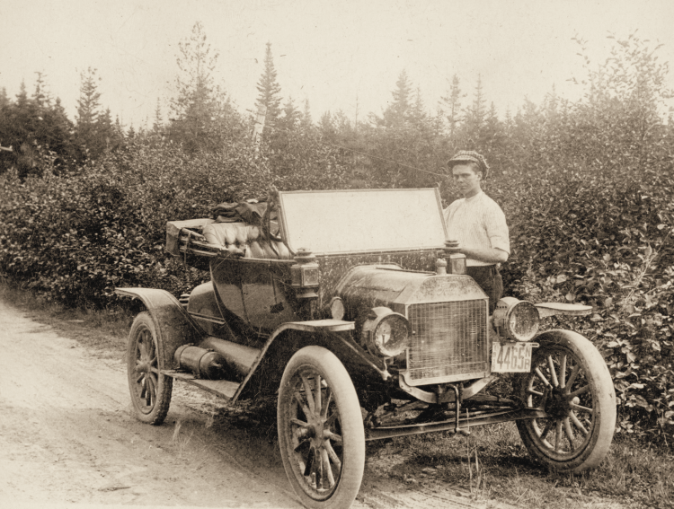 Cub, wearing a cap, stands beside the Keith family’s open-top car on a road bordered by shrubs and trees.