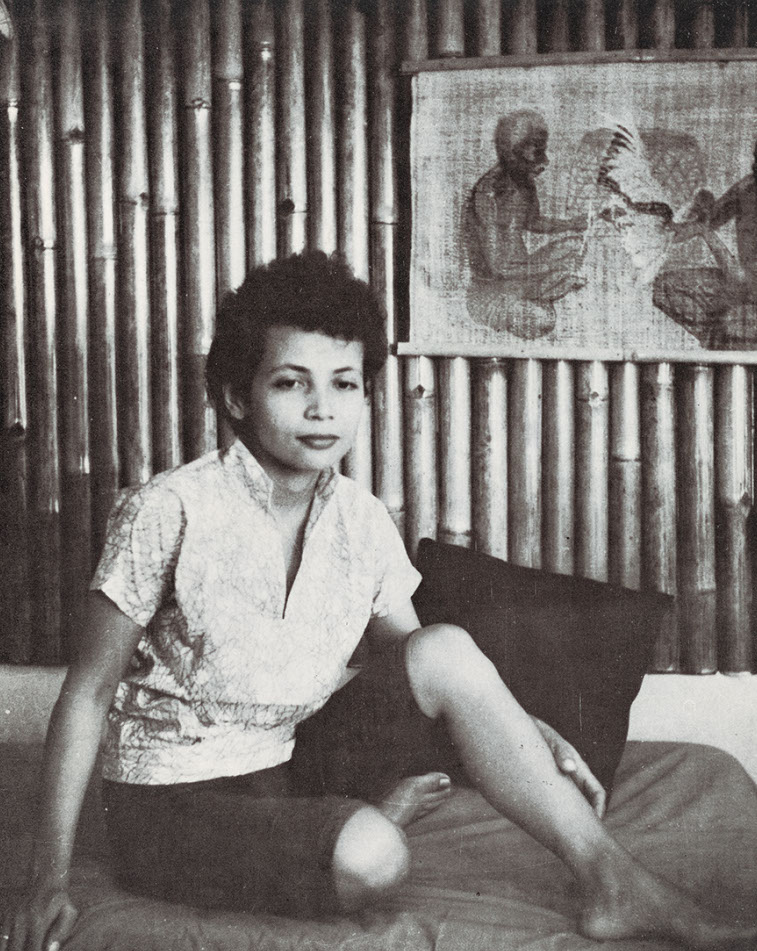 Ger sits on a bed with one knee raised, wearing a light patterned blouse and a dark skirt; a framed picture hangs behind her.