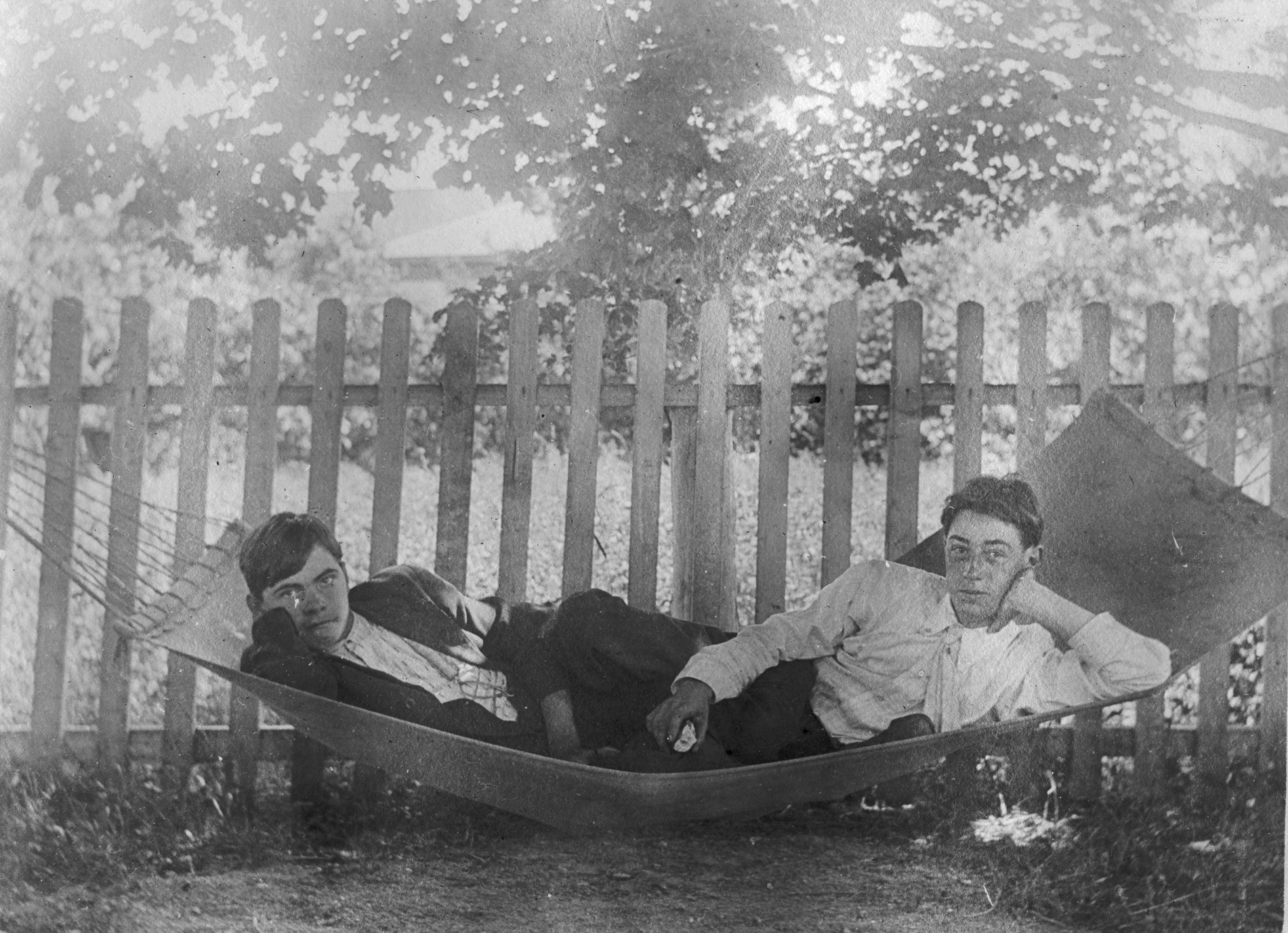 Len and Cub recline at opposite ends of a low hammock beneath leafy branches, with a picket fence behind them.