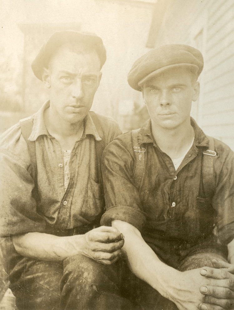 Len and Cub sit side by side in caps, suspenders and marked work clothes; one holds the other’s forearm near the elbow.