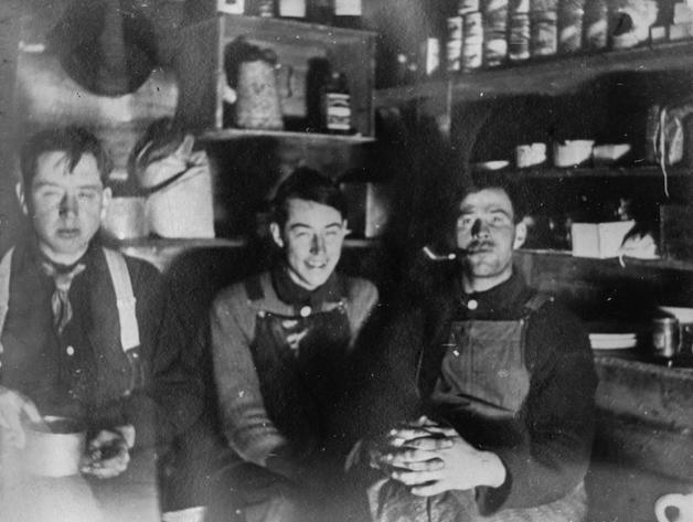 Len sits between his brother Frank and Roy Corey in a room lined with shelves of tins, with a cup and pipe visible.